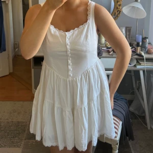 Vit ärmlös klänning med knappar - Säljer en söt vit klänning med knappar framtill och spetsdetaljer vid axelbanden. Klänningen har en luftig passform och volangdetaljer i kjolen. Perfekt för varma sommardagar!