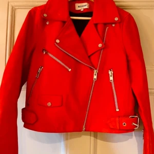 Röd bikerjacka från Weekday - Snygg röd bikerjacka från Weekday med dragkedjor och knappar i silver. Jackan har en klassisk bikerstil med asymmetrisk dragkedja och bälte i nederkant. Perfekt för att ge din outfit en cool touch. Äkta skinn ock mocka storlek 38.
