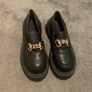 Svarta loafers med guldkedja - Snygga svarta loafers i skinn med en dekorativ guldkedja på ovansidan. Skorna har en robust sula som ger bra grepp. Perfekta för en stilren look.💗 helt nya, aldrig använda!