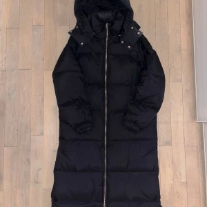 Svart lång dunjacka med huva ARKET - Säljer en svart lång dunjacka med huva och dragkedja. Jackan har långa ärmar och är perfekt för kalla dagar. Den har en stilren design med tryckknappar vid kragen. Tecken på användning