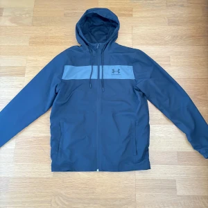 Svart vindjacka från Under Armour - Säljer en stilren svart vindjacka från Under Armour med grått band över bröstet och justerbar huva. Jackan har dragkedja framtill och två sidofickor. Perfekt för träning eller vardagsbruk. Vid snabb affär kan pris diskuteras 