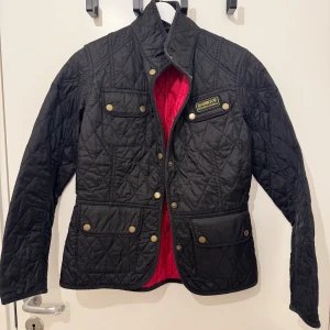 Svart quiltad jacka från Barbour - Säljer en snygg svart quiltad jacka från Barbour med röd insida. Jackan har en klassisk design med guldknappar och en Barbour International-logga på bröstet. Perfekt för en stilren look!