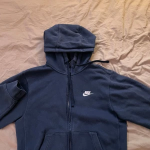 Mörkblå hoodie från Nike - Säljer en mörkblå hoodie från Nike med dragkedja och klassisk logga på bröstet. Perfekt för en avslappnad stil. Den har en justerbar huva och fickor framtill.