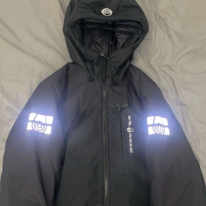 Svart jacka från Sail Racing - Säljer en svart jacka från Sail Racing med reflekterande detaljer på ärmarna och bröstet. Jackan har en huva och dragkedja framtill. Perfekt för kyliga dagar. Priset kan sänkas 