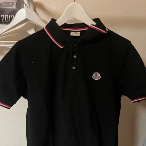 Svart pikétröja från Moncler - Snygg svart pikétröja från Moncler med klassisk krage och knappar framtill. Tröjan har röda och vita detaljer på krage och ärmslut samt Monclers logga på bröstet. Perfekt för en stilren look.