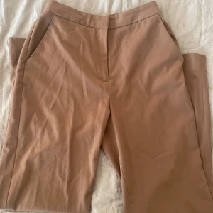 Bruna kostymbyxor - Snygga bruna kostymbyxor med hög midja och raka ben. Byxorna har diskreta fickor både fram och bak samt stängs med dragkedja och knapp. Perfekta för en stilren look.
