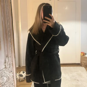 Svart kappa med vita detaljer - Snygg svart kappa med vita broderade detaljer längs kanterna. Jackan har en omlott-design och knyts med ett bälte i midjan. Perfekt för att ge en stilren look.
