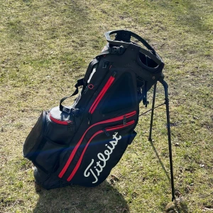 Svart och röd golfbag från Titleist - Säljer en stilren och praktisk golfbag från Titleist i svart med röda detaljer. Bögen, har använts i en golf säsong så är typ helt ny. Bagen har flera fack med dragkedjor och en stabil ställning för enkel användning på golfbanan. nypris på vägen är runt 4000