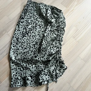 Mönstrad kjol med knyt - Snygg kjol med svart leopardmönster på ljus bakgrund. Kjolen har en knytning på sidan som ger en fin detalj och volangkant nedtill. Perfekt för att ge en trendig touch till din outfit.
