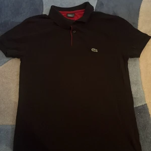 Svart pikétröja från Lacoste - Säljer en stilren svart pikétröja från Lacoste med klassisk krage och knappdetalj. Tröjan har en liten grön krokodil-logga på bröstet och röd insida vid kragen. Perfekt för en avslappnad men snygg look.