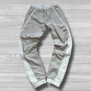 Adidas byxor - Säljer ett par grå träningsbyxor från Adidas med elastisk midja och muddar vid bensluten. Byxorna har en vit rand längs sidorna och en liten Adidas-logga på benet. Perfekta för en sportig look!