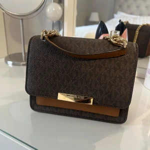 Brun axelväska från Michael Kors - Snygg brun axelväska från Michael Kors med guldfärgade kedjedetaljer och logotyp på framsidan. Väskan har ett elegant mönster och en praktisk storlek för alla dina nödvändigheter.