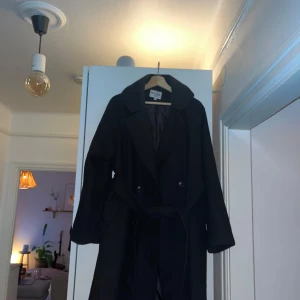 Svart kappa med bälte NA-KD - Elegant svart kappa med bälte och långa ärmar. Perfekt för kyligare dagar och ger en stilren look. Passar bra till både vardag och mer formella tillfällen.