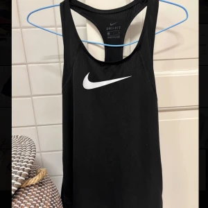 Svart linnetopp från Nike - Snygg och sportig svart linnetopp från Nike med Dri-FIT-teknologi. Perfekt för träning med sin lätta och andningsbara design. Den har en klassisk rund halsringning och ett stilrent vitt Nike-logotyptryck på framsidan🏃‍♀️