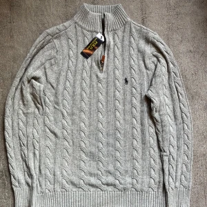 Grå stickad tröja från Ralph Lauren LÄS BESKRIVNINGEN - OBS! XL men passar bäst om du är L. Säljer en stilren grå stickad tröja från Ralph Lauren med dragkedja och kabelstickat mönster. Tröjan har långa ärmar och en liten broderad logga på bröstet. Perfekt för kyliga dagar!