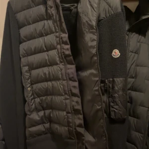 Moncler Cardigan  - Säljer en snygg svart jacka från Moncler med quiltad design och stickade ärmar. Jackan har en huva och dragkedja framtill samt en praktisk ficka med dragkedja på ärmen. Perfekt nu för våren/sommaren! Den är storlek XL men passar även L