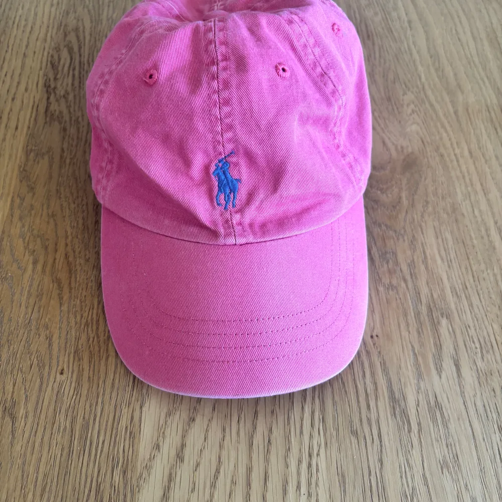 Snygg rosa keps från Polo Ralph Lauren med logga i blått framtill. Justerbar rem baktill för perfekt passform. Perfekt för en avslappnad stil.. Asusteet.