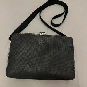 Svart axelväska Lacoste - Hej! Säljer nu min helt nya Lacoste väska då den aldrig kommit till användning.  Där är använt 1 gång och är sprillans ny. Köpt för 2200kr på Lacoste officiella hemsida jag säljer för 1400kr men pris kan diskuteras vid snabb affär. 