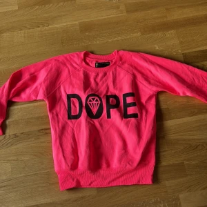 Rosa sweatshirt från Brave New World - Säljer en cool rosa sweatshirt från Brave New World med texten 'DOPE' och en diamantgrafik i svart på framsidan. Tröjan har långa ärmar och en avslappnad passform. Perfekt för en avslappnad stil!