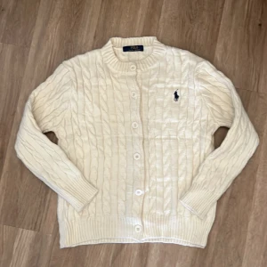 Vit stickad kofta från Ralph Lauren - Säljer en stilren vit stickad kofta från Ralph Lauren med kabelstickat mönster och knappar framtill. Den har långa ärmar och en broderad logotyp på bröstet. Perfekt för kyliga dagar!