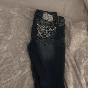 Mörkblå jeans med broderade fickor från Miss Me - Snygga mörkblå jeans från Miss Me med unika broderade detaljer och strass på bakfickorna. Jeansen har en medelhög midja och en klassisk femficksdesign. Perfekta för att ge din outfit en extra touch av stil. Dem är mid waisted jag säljer dm för att de är förstora.💕dom är lite sönder trampade💕