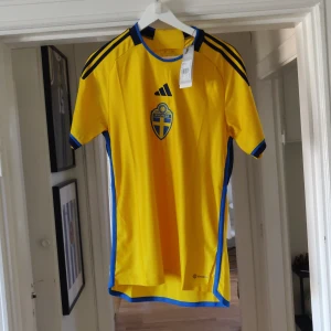 Sverige fotbollströja från Adidas - Säljer en gul fotbollströja från Adidas med blå detaljer och Sveriges landslagsemblem på bröstet. Tröjan har korta ärmar och en modern passform. Perfekt för fotbollsfans!köpt ifrån adidas, etikett finns kvar och aldrig använd. Finns på fler sidor, vid frågor eller fler bilder är det bara att skriva👍