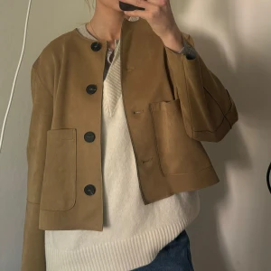 Beige mockajacka med stora knappar - Säljer en beige mockajacka med stora svarta knappar och två framfickor. Jackan har en kortare passform och är långärmad. 