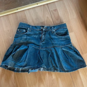 Jeanskjol med volang - Säljer en blå jeanskjol med volang och klassiska fickor fram och bak. Kjolen har en kort passform och stängs med knapp och dragkedja framtill. Perfekt inför sommaren. Midjemåttet rakt över är 37, men den är insydd så går att få större. 