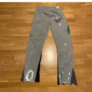 Gallery Dept Painted Flare Sweat Pants  - Grå mjukisbyxor från Gallery Dept. med utsvängda ben och färgstänk i vitt och grönt. Byxorna har elastisk midja och tryckt logga på ena låret. Unik design med konstnärlig känsla och råa benslut. Äkta från stockX 