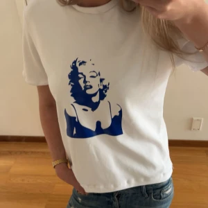 Vit t-shirt med blått marilyn monroe tryck - Vit t-shirt med ett stort blått tryck av Marilyn Monroe på framsidan och texten 'GOOD SHOES TAKE YOU TO GOOD PLACES' på ryggen. Sytt den själv därav priset, klassisk passform och rund halsringning, perfekt till jeans.