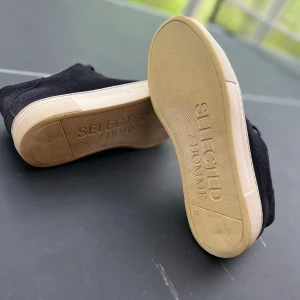 Svarta sneakers från Selected Homme - Säljer ett par svarta sneakers från Selected Homme med beige sula och snörning. Skorna har en stilren design och är tillverkade i mocka med en mjuk insida. Perfekta för dig som gillar klassiska och bekväma skor.