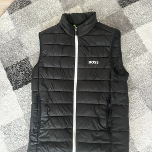 Svart dunväst från BOSS - Stilren svart dunväst från BOSS med vit dragkedja framtill och diskret logga på bröstet. Västen har ståkrage och två sidofickor med dragkedja. Perfekt för lager-på-lager under kyliga dagar.