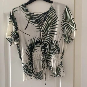 H&m blus - Säljer en blus med  palmbladsmönster i grönt och vitt. Blusen har korta ärmar, v-ringning och knytning framtill samt resår i midjan för en snygg passform. Bra skick storlek S