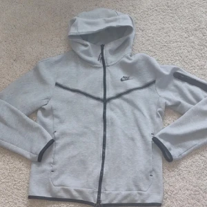 Nike tech - Snygg grå hoodiejacka från Nike med svart dragkedja och svarta detaljer. Jackan har huva, lång ärm och två fickor framtill. Perfekt för en sportig och avslappnad stil.