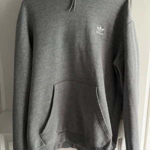 Grå hoodie från Adidas - Säljer en klassisk grå hoodie från Adidas med vit logga på bröstet. Tröjan har huva med dragsko och en stor magficka framtill. Perfekt för en avslappnad stil och enkel att matcha med det mesta.