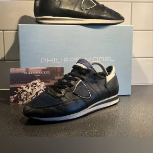 Svarta sneakers från Philippe Model - Säljer ett par svarta Philippe Model sneakers med detaljer i mörkblått tyg och vita inslag vid hälen. Skorna har snörning och en platt sula, samt en sportig design som passar till många outfits. Säljer åt en kompis men tveka inte på att höra av er om frågor