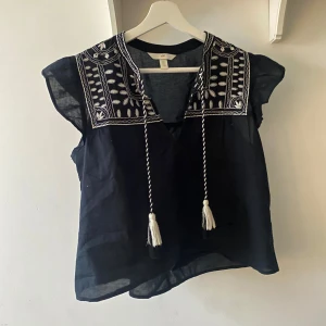 Svart blus med broderade detaljer och tofsar - Svart blus med vita broderade mönster på axlarna och bröstet. Perfekt för en avslappnad men ändå stilren outfit😍 
