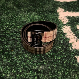 Beige och brun mönstrat bälte med metallspänne - Säljer ett snyggt bälte med klassiskt rutigt mönster i beige och bruna toner. Bältet har ett stort metallspänne med BT logotyp i svart. Perfekt accessoar för att lyfta din outfit. Vid mer funderingar skriv!!! Priset går att diskutera 