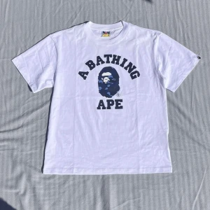 Vit t-shirt från A Bathing Ape - Säljer en klassisk vit t-shirt från A Bathing Ape med stort tryck i blått och svart på bröstet. T-shirten har rund hals och korta ärmar. Ikonisk BAPE-logga på ärmen. Perfekt för dig som gillar streetwear.