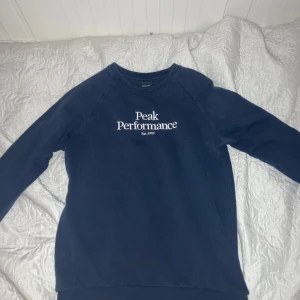 Mörkblå sweatshirt från Peak Performance - Säljer en mörkblå sweatshirt från Peak Performance med vit logotyp på bröstet. Tröjan har rund halsringning och långa ärmar. Perfekt för en avslappnad stil.