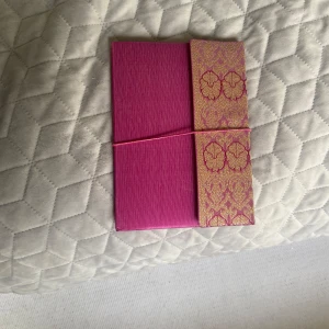 Rosa anteckningsbok med guldmönster - Säljer en elegant oanvänd anteckningsbok i stark rosa färg med ett dekorativt guldmönstrat band på framsidan. Boken har tomma vita sidor och ett elastiskt band för att hålla den stängd. Perfekt för anteckningar, dagbok eller skisser.hämtas i norsborg botkyrka eller fraktas.