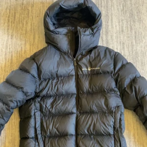 Svart dunjacka från Sail Racing - Säljer en svart dunjacka från Sail Racing med huva och dragkedja. Jackan är quiltad och har en klassisk pufferstil som håller dig varm under kalla dagar. Perfekt för dig som vill ha en stilren och funktionell jacka.