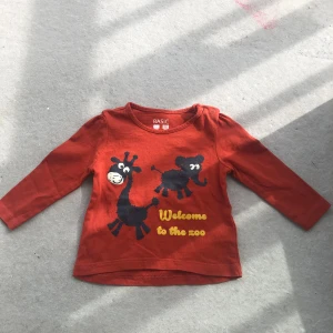Rostfärgad långärmad tröja med djurtryck - Säljer en söt rostfärgad långärmad tröja från H&M Basic med tryck av giraff och elefant samt texten 'Welcome to the zoo' i gult. Tröjan har knappar vid axeln för enkel påklädning. Perfekt för dig som gillar färg och lekfulla motiv!
