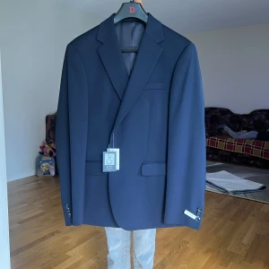 Mörkblå kavaj från Oscar Jacobson - Säljer en stilren mörkblå kavaj från dressman. Klassisk modell med två knappar framtill, smala slag och fickor med lock. Perfekt för formella tillfällen eller bal. Kavajen är i ett mjukt material och har en elegant passform.