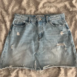 Ljusblå jeanskjol från Zara - Säljer en ljusblå jeanskjol från Zara med slitna detaljer och rå kant nertill. Kjolen har klassisk femficksmodell och knappgylf. Perfekt för en avslappnad och trendig look.
