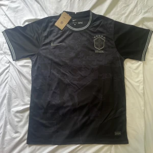 Svart Brasilien fotbollströja från Nike - Säljer en svart Brasilien fotbollströja från Nike med diskret camomönster och broderat klubbmärke på bröstet. Tröjan har korta ärmar och ribbade detaljer vid hals och ärmslut. Perfekt för dig som vill sticka ut på planen.
