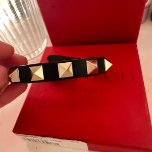 Svart läderarmband med nitar från Valentino - Snyggt svart armband i läder från Valentino med stora fyrkantiga guldfärgade nitar och spänne i metall. Kommer i originalask med tygpåse. Perfekt accessoar för att lyfta din stil.