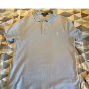 Ljusblå pikétröja från Polo Ralph Lauren - Säljer en klassisk ljusblå pikétröja från Polo Ralph Lauren med korta ärmar och krage. Tröjan har knappar framtill och den ikoniska broderade loggan på bröstet. Perfekt för en stilren och avslappnad look. Pris kan diskuteras vid snabb affär