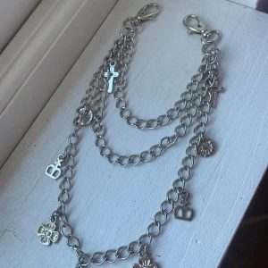 Silverfärgat kedjebälte med berlocker - Ett silverfärgat kedjebälte med flera hängen i form av blommor, kors och bokstäver. Bältet har tre kedjor i olika längder och stora knäppen för att enkelt knäppa fast på dina byxor. Perfekt accessoar för att toucha upp din outfit!