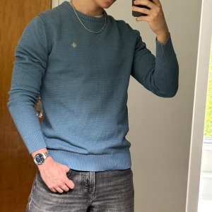 Blå stickad tröja från Morris - Säljer en stilren blå stickad tröja från Morris. Tröjan har rund halsringning, diskret logga på bröstet och ribbade muddar vid ärmslut och nederkant. Perfekt för en snygg och avslappnad look. Tröjan är i m men sitter som en s 